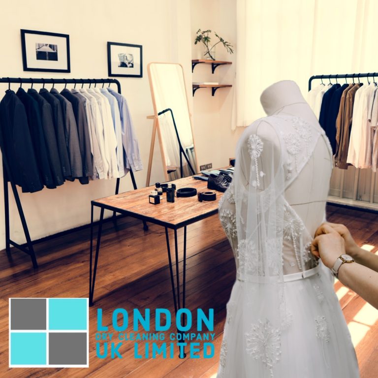 A bridal gown being adjusted on a mannequin in a stylish room with racks of suits and a wooden table. The room has a warm, elegant ambiance. Logo at the bottom reads London Dry Cleaning Company UK Limited.