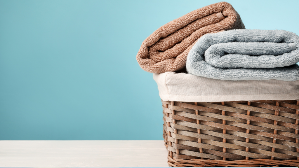 A wicker basket sits on a white surface against a blue background, holding two neatly folded towels—one brown and one gray, fresh from London Dry Cleaning.