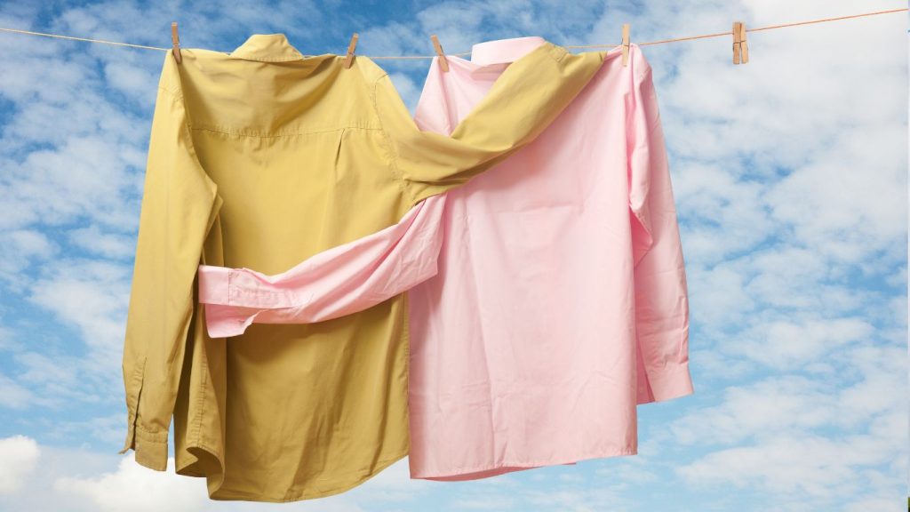 Two long-sleeve shirts, one yellow and one pink, are hanging on a clothesline against a bright blue sky with scattered white clouds, freshly laundered to perfection by London Dry Cleaning.